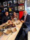 Two men and a woman at a table with food in the middle of them. 